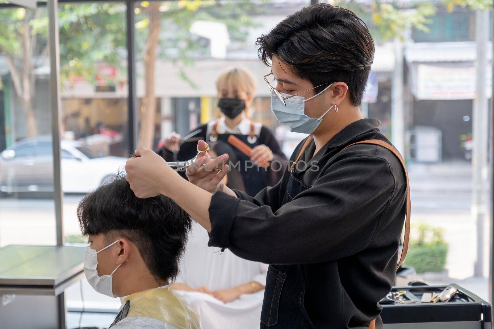 Beauty salon Asian barber man with hygiene mask use scissors cut hair of customer in the shop with other co-worker also take care hair style of woman. Beauty business for good appearance concept. by nrradmin