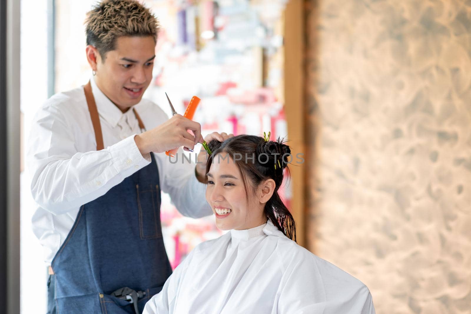 Beautiful Asian woman sit and smile in beauty salon shop that take care of hair cut by barber man. They look happy and enjoy during the process with beauty business concept.