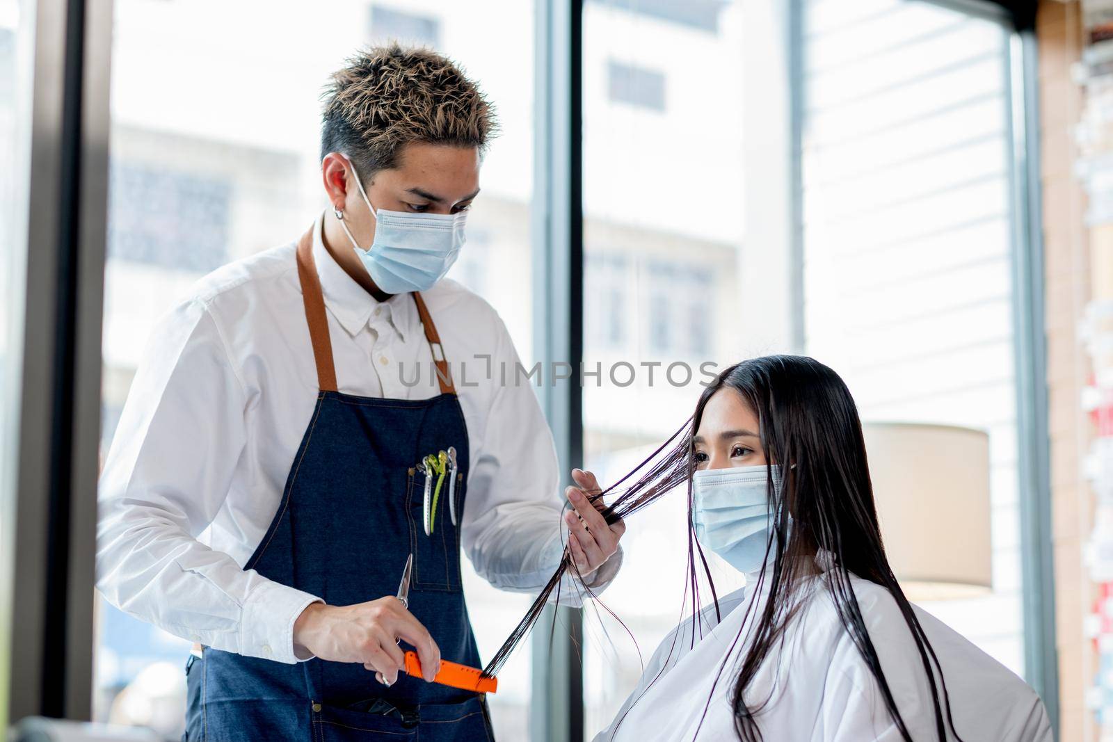 Asian beauty salon barber man with hygiene mask stand and process of hair cut of long hair customer and woman also wear hygiene mask. Concept of beauty business during Covid-19 pandemic.