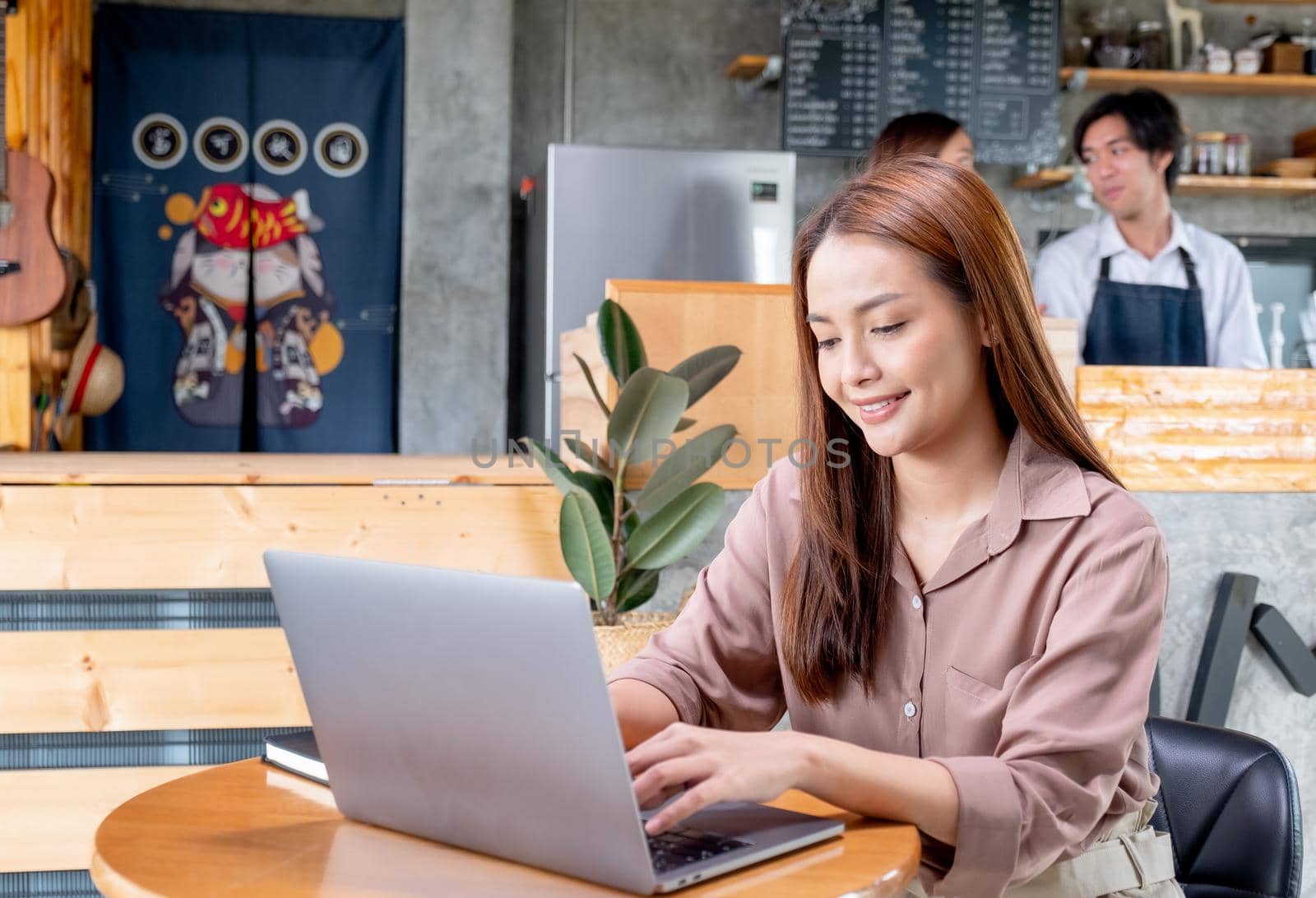 Beautiful Asian woman work with laptop in coffee shop and two barista or coffee maker work together in the back. Concept of small business and support by customer.