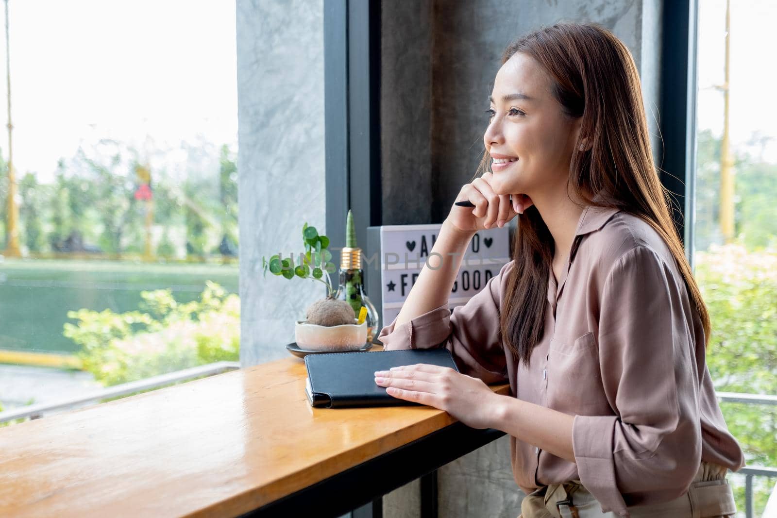 Beautiful Asian woman sit in corner of coffee shop with day light and look outside. She is smiling with happy emotion and think about her small business and sustainable activity.