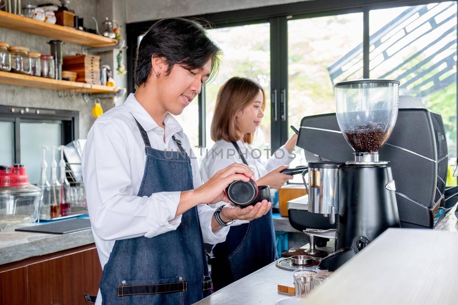 Barista man or coffee maker pour cream or ingredient to the cup and stand beside of co-worker man in coffee shop. Concept of happy working with small business and sustainable together.