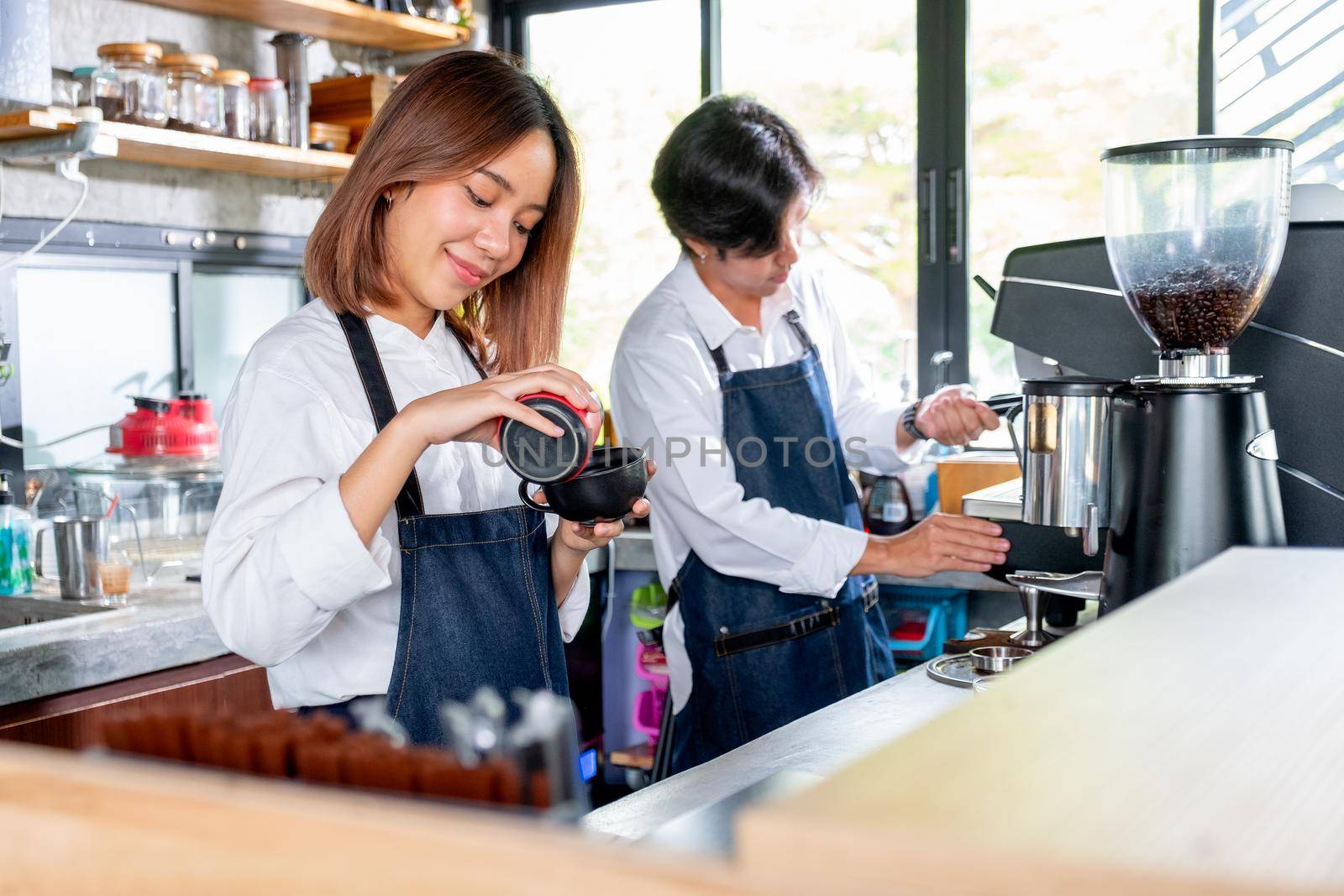 Barista woman or coffee maker pour cream or ingredient to the cup and stand beside of co-worker man in coffee shop. Concept of happy working with small business and sustainable together.