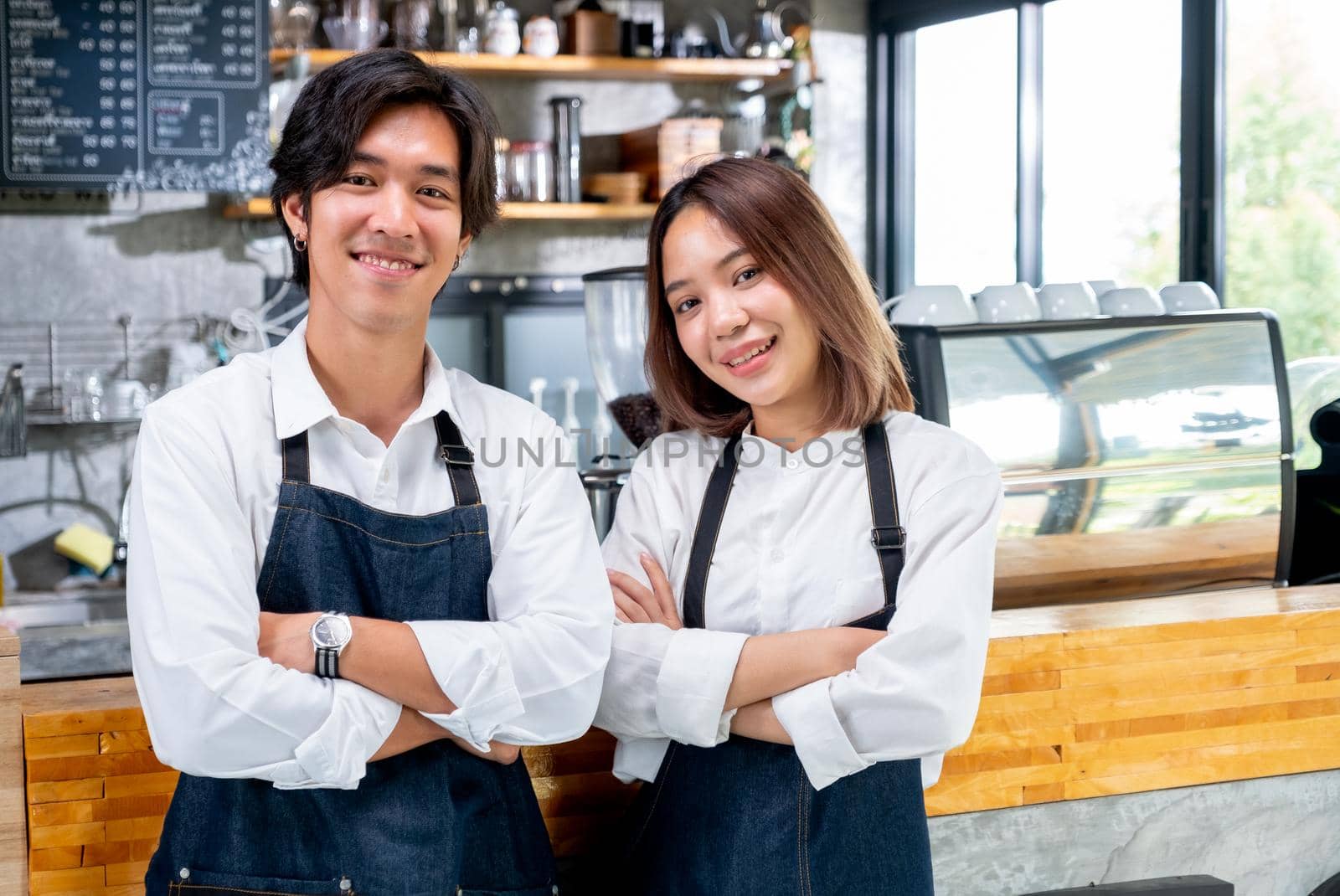 Two Asian barista or coffee maker man and woman stand with arm-crossed or confidence action also look to camera and smile in coffee shop. Concept of happy working with small business together.