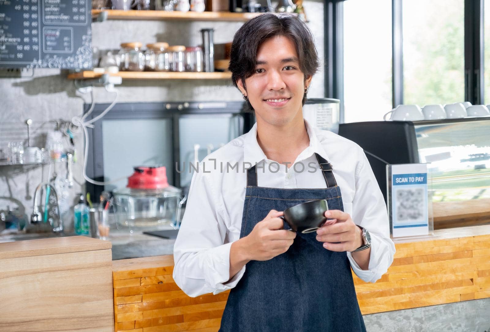 Barista man of coffee maker hold a cup and stand in front of counter in coffee shop with looking at camera and smile. Concept of happy working with small business and sustainable.