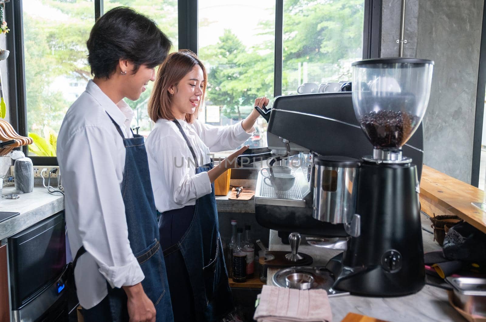 Barista man stand near his co-worker who prepare of produce coffee using the coffee maker machine and they look happy by smiling for working together.