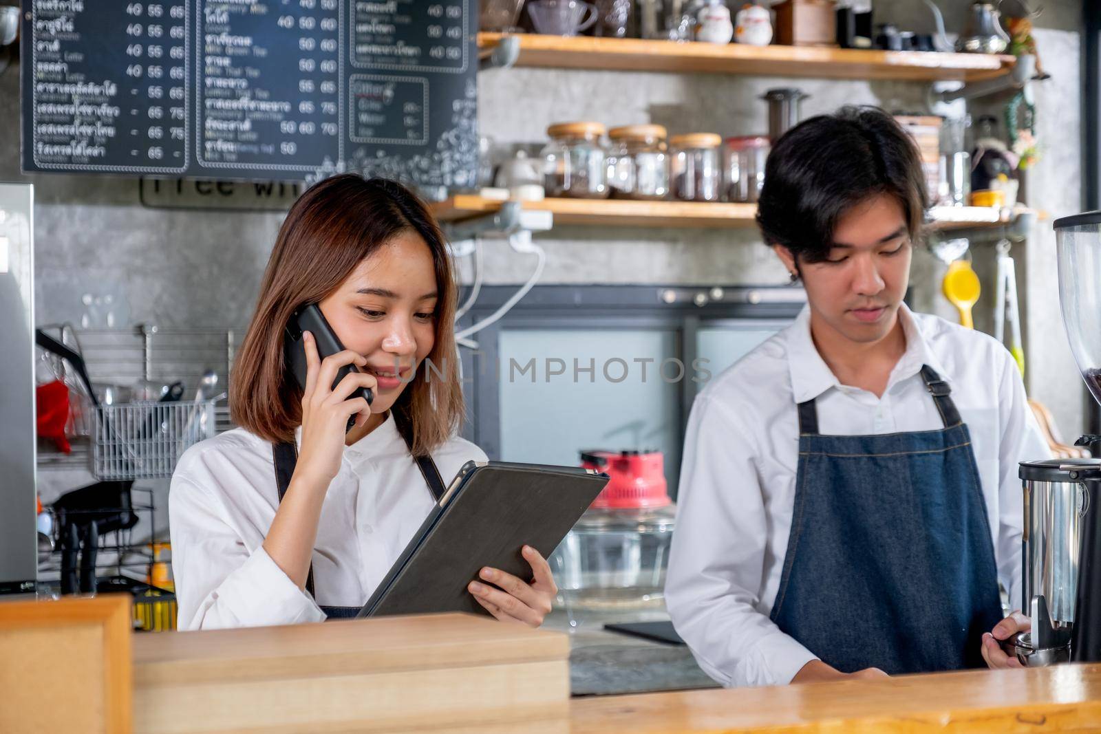 Barista girl or coffee maker receive phone call and record order and stand beside of co-worker man in coffee shop. Concept of happy working with small business and sustainable together.