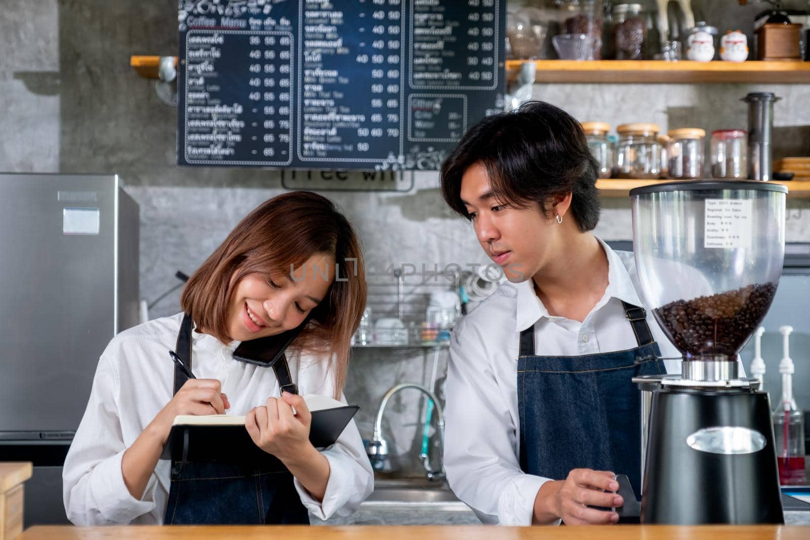 Barista girl or coffee maker answer mobile phone also record the order detail into notebook while her co-worker is working with machine. Concept of good teamwork support small business system.