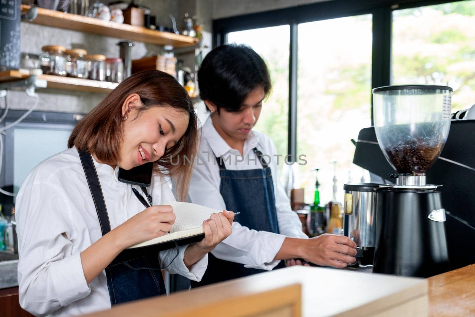 Barista girl or coffee maker answer mobile phone also record the order detail into notebook while her co-worker is working with machine. Concept of good teamwork support small business system.