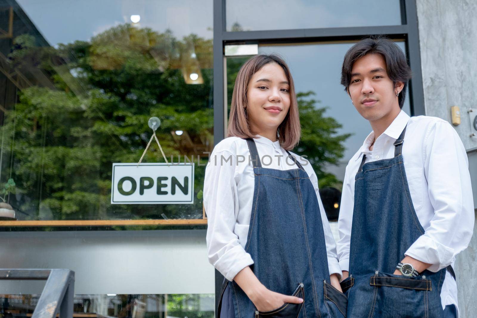 Two Asian barista or coffee maker man and woman stand with confidence action also look at camera and smile in front of coffee shop with day light.