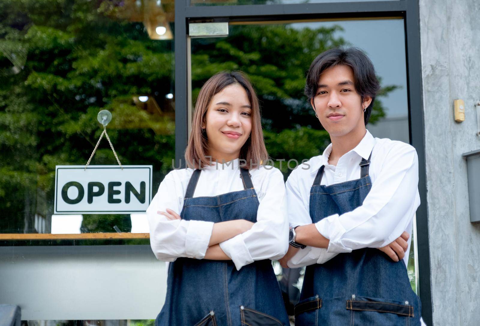 Two Asian barista or coffee maker man and woman stand with arm-crossed or confidence action also look to camera and smile in front of coffee shop. Concept of happy working with small business.