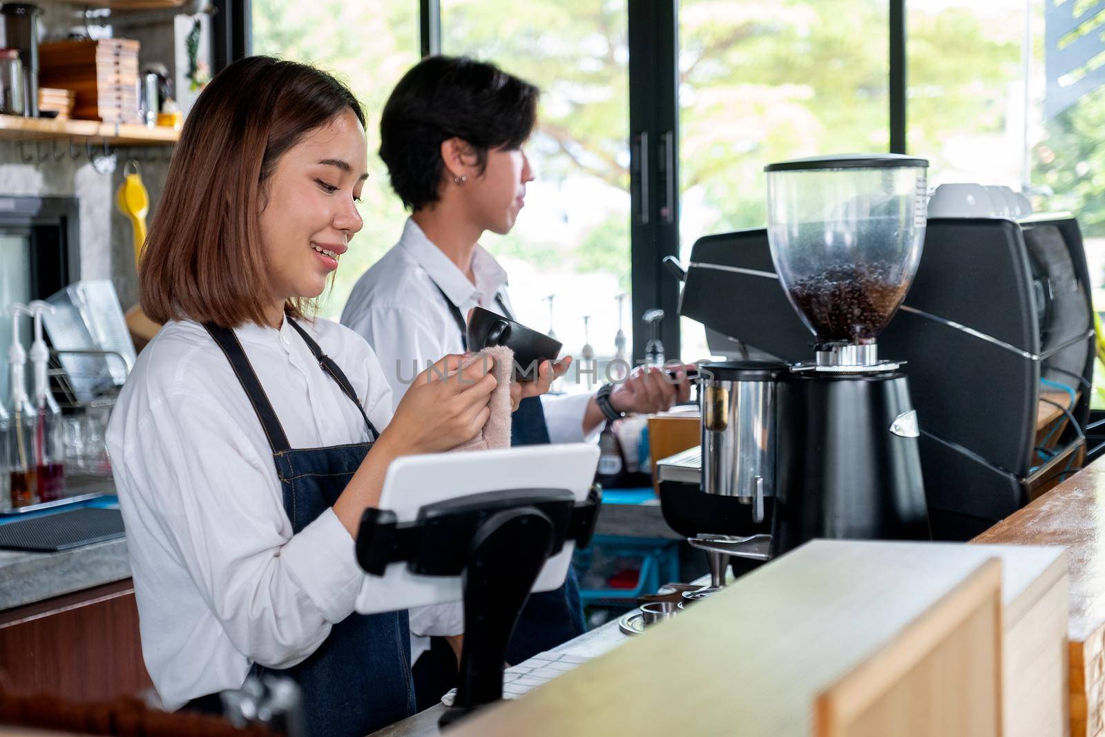 Asian barista or coffee maker woman clean cup of coffee while her co-worker man work with machine in coffee shop. Concept of happy working with small business together.