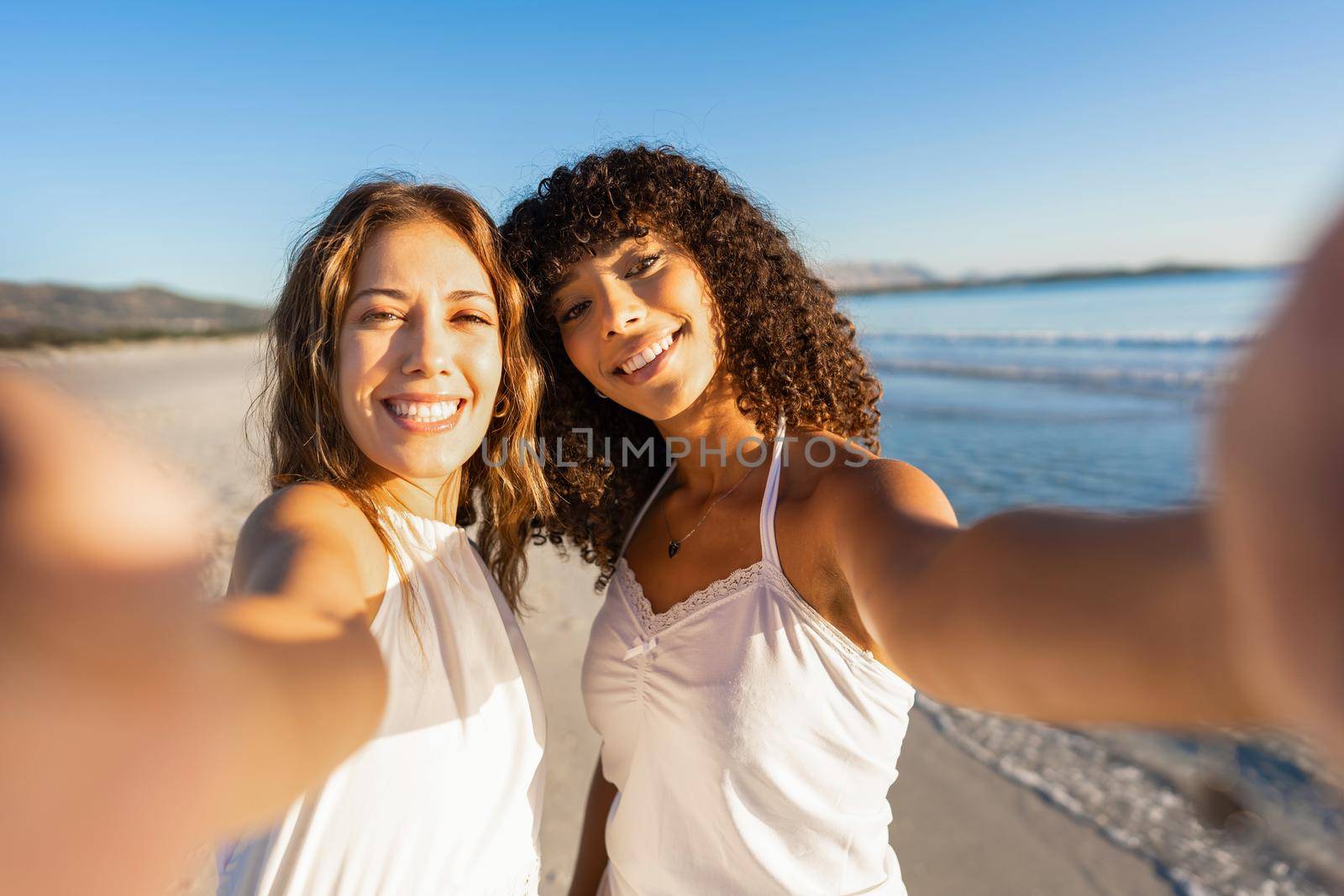 Two beautiful mixed race girl holding smartphone making a self portrait on beach smiling at camera wearing boho white dress at sunset or dawn. Gay couple of young millennial women in tropical vacation by robbyfontanesi