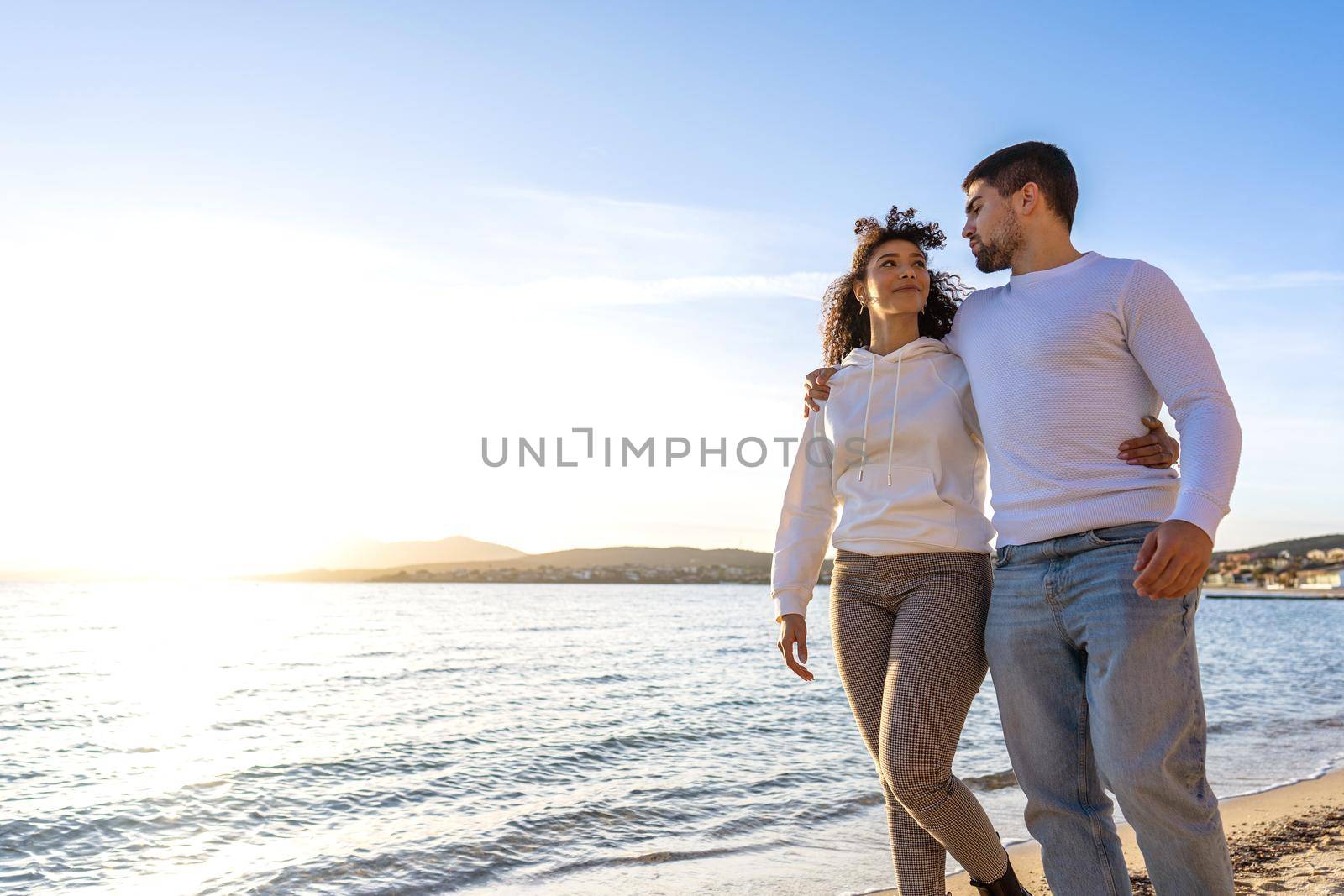 Multiracial couple of young millennials in love embracing each other while walking by the sea at sunset or sunrise looking into each other's eyes. Concept: affections has no boundaries of race or sex