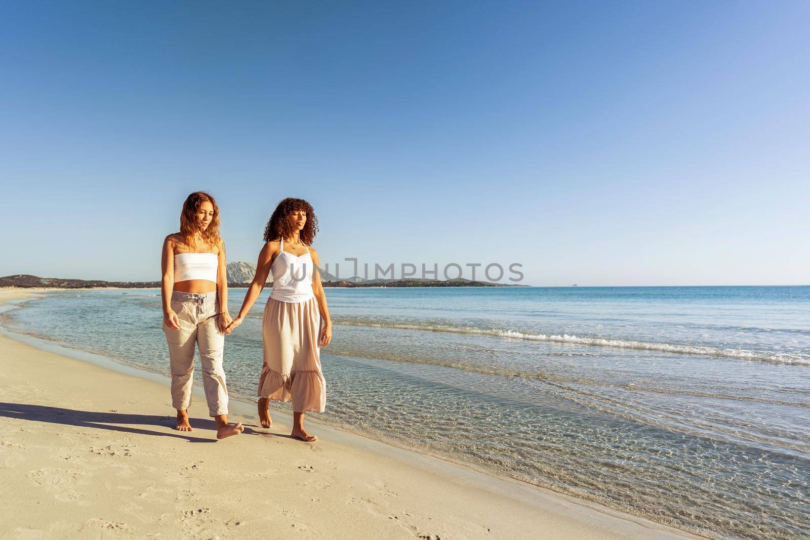 Multiracial couple of beautiful young gay women holding hands walking at sunrise or sunset on a tropical colored beach with flat ocean sea water. Concept: in love and nature there are no differences