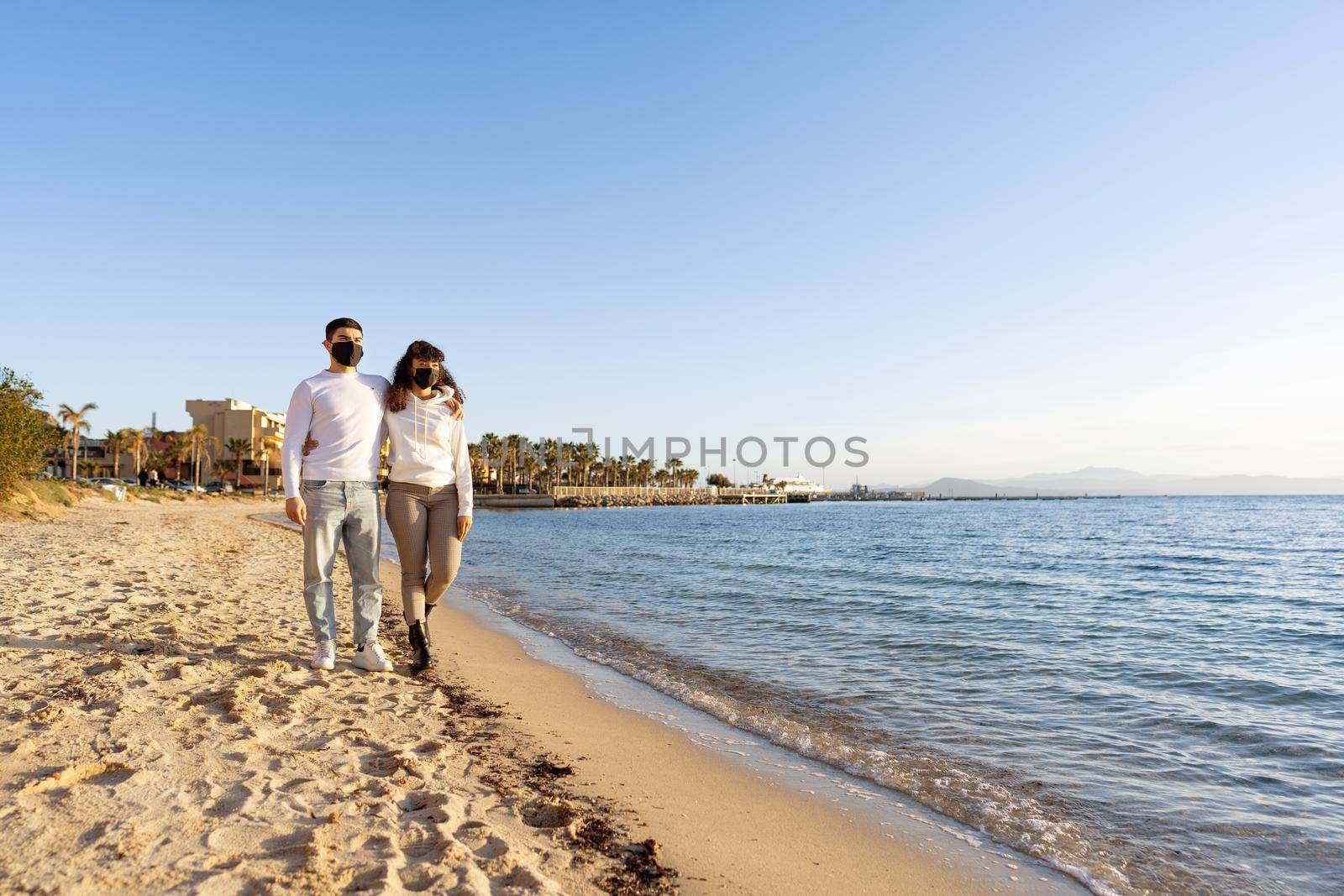 Multiracial couple of young millennials in love embracing each other while walking by the ocean seaside at sunset or sunrise wearing protective face mask against Coronavirus in new normal vacations