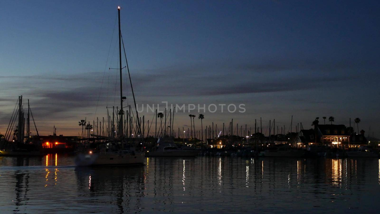 Luxury yachts sailboats floating, marina harbour quay. Sail boat masts, nautical vessels in port. Harbor fisherman village in Oceanside, California USA. Evening dusk, twilight lights and palm trees.