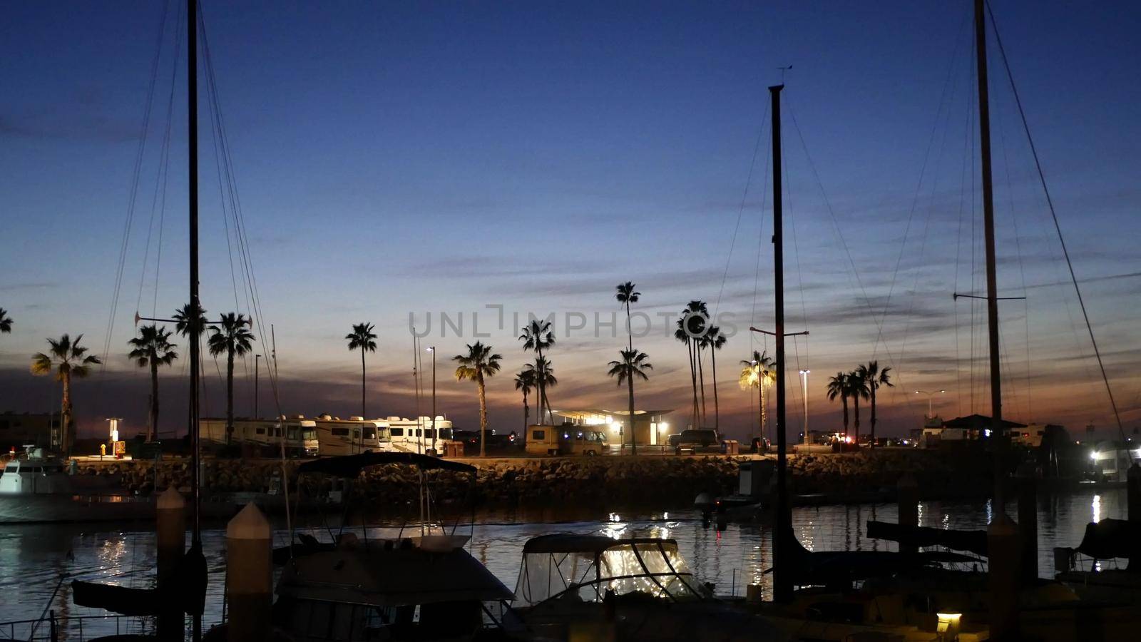 Luxury yachts sailboats floating, marina harbour quay. Sail boat masts, nautical vessels in port. Harbor fisherman village in Oceanside, California USA. Evening dusk, twilight lights and palm trees.