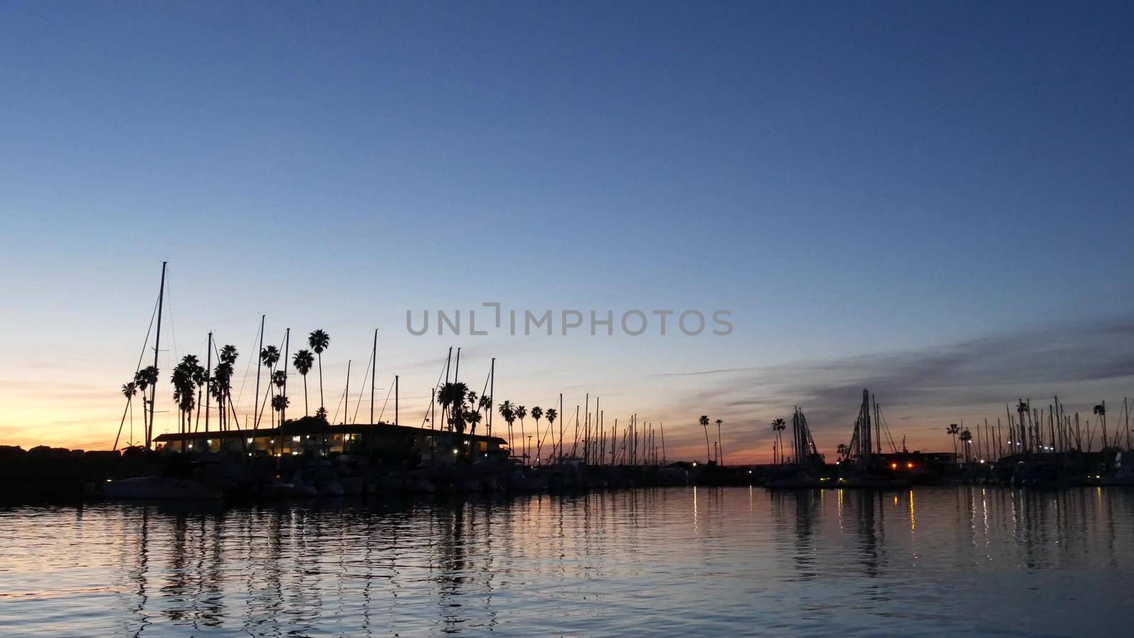 Luxury yachts sailboats floating, marina harbour quay. Sail boat masts, nautical vessels in port. Harbor fisherman village in Oceanside, California USA. Evening dusk, twilight lights and palm trees.