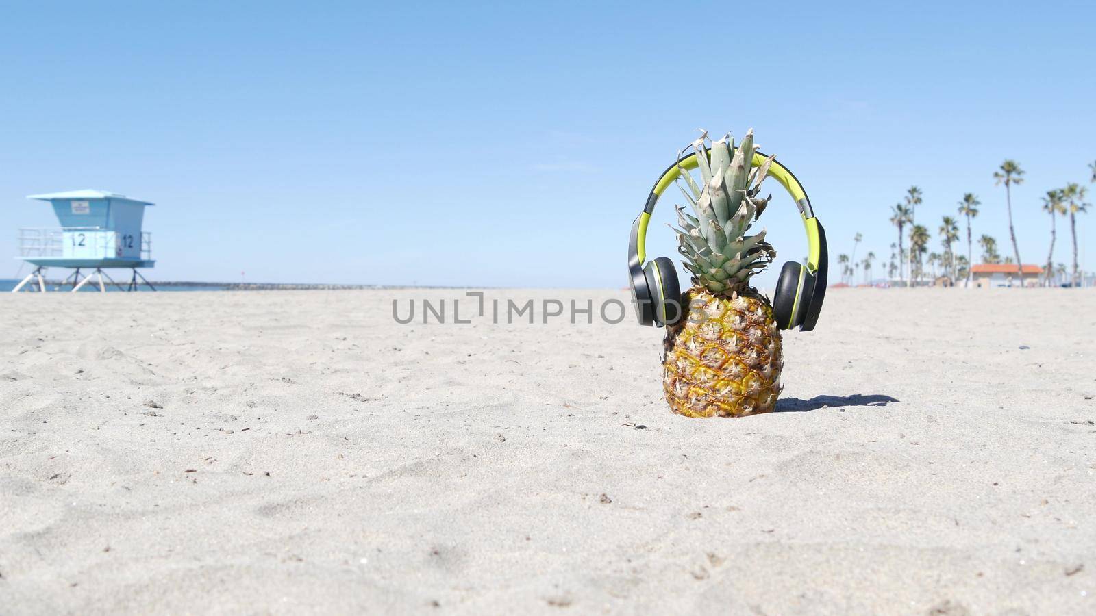 Funny pineapple in headphones, sandy ocean beach by blue lifeguard hut, California USA, Pacific coast. Tropical summer exotic fruit enjoying vacations and music in paradise. Ananas sunbathing on shore