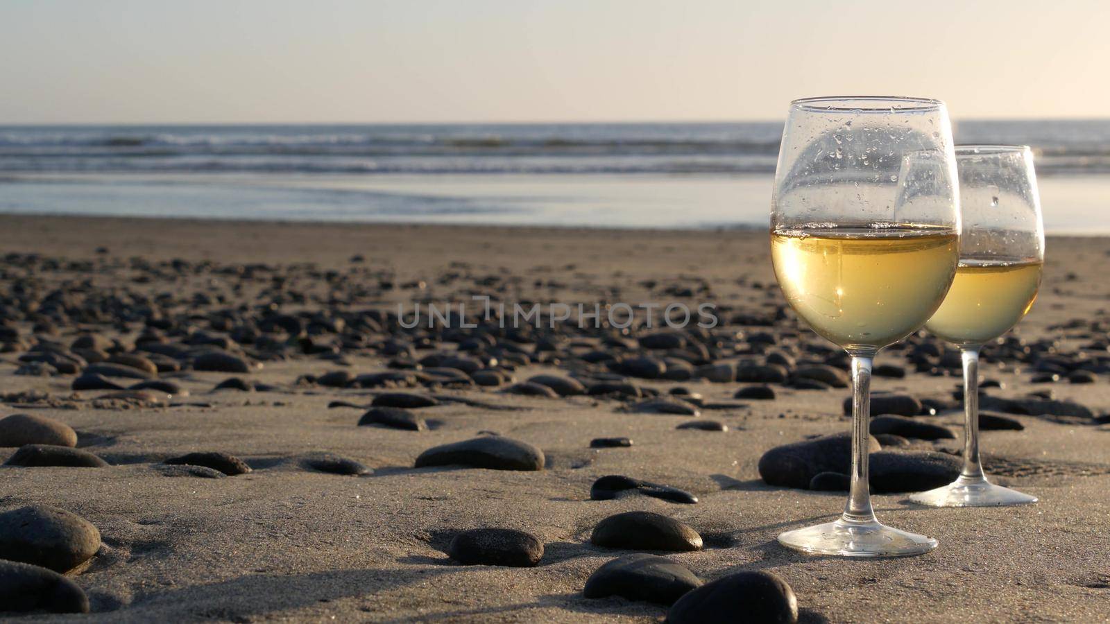 Two wineglasses on sandy ocean beach. Couple of glasses and pebbles, white wine for romantic date near sea water. Cozy beachfront sunset near tide waves. Seamless looped cinemagraph. California, USA.