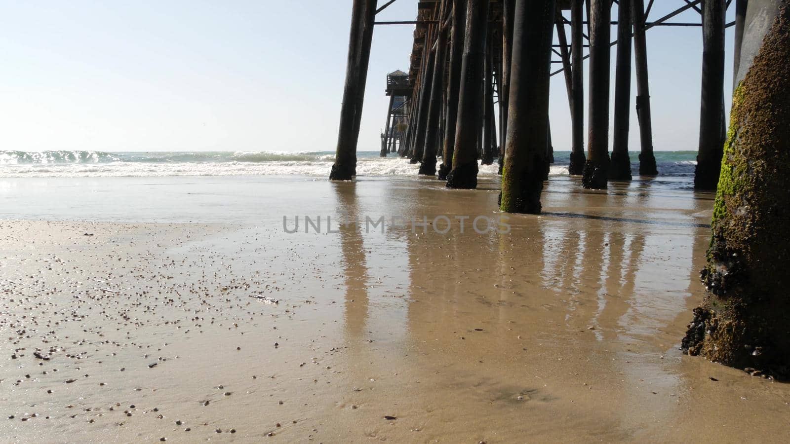 Wooden piles under boardwalk, old pier in Oceanside, California coast USA. Pilings, pylons or pillars below retro vintage bridge, waterfront promenade. Ocean waves, sea water tide and sand beach.