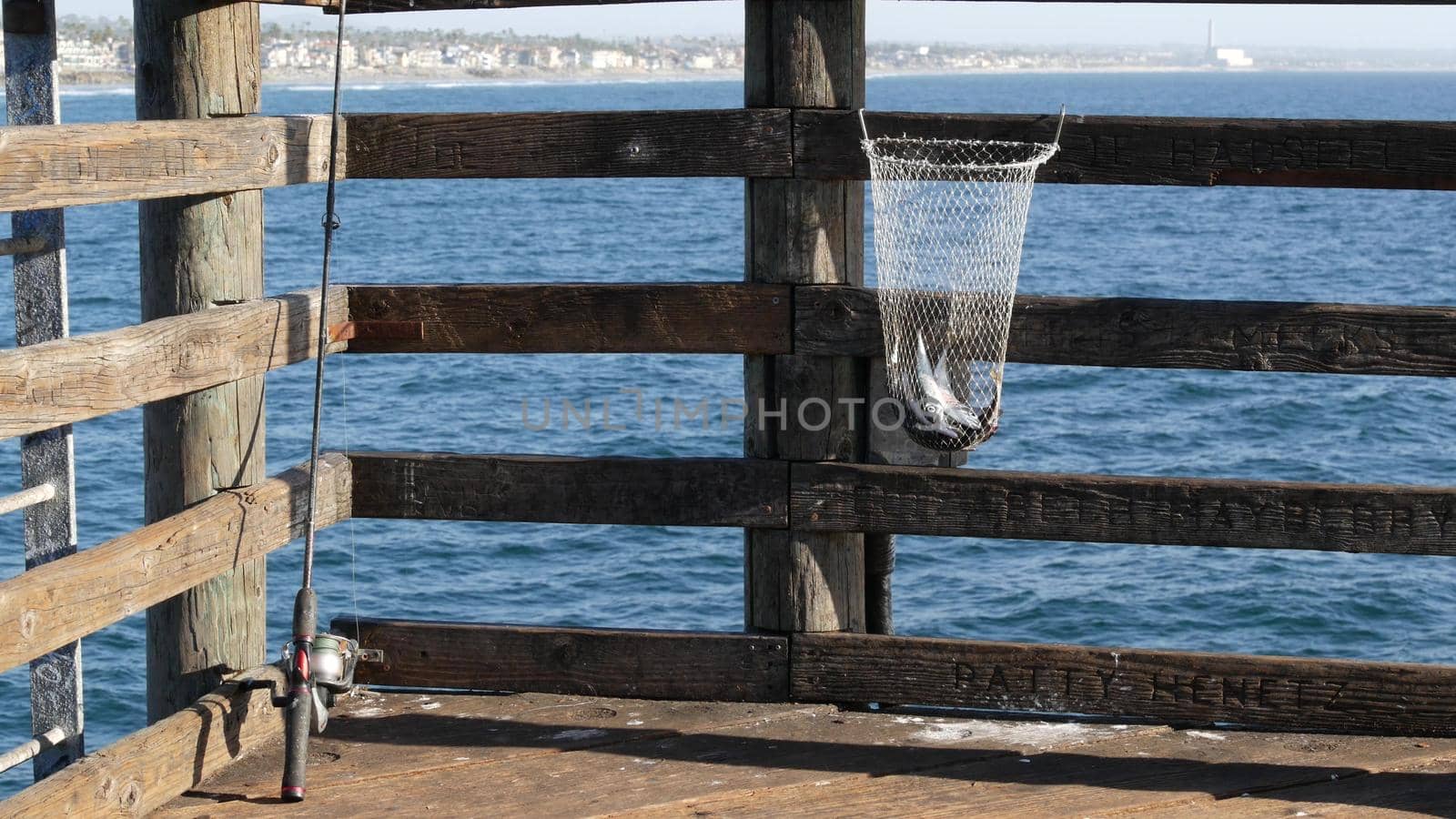 Saltwater angling, wooden pier boardwalk, fishing accessory, tackle or gear. Oceanside California USA. Sea salt water, ocean seascape. Rod or spinning, fisherman catch, fresh alive fish in net basket.