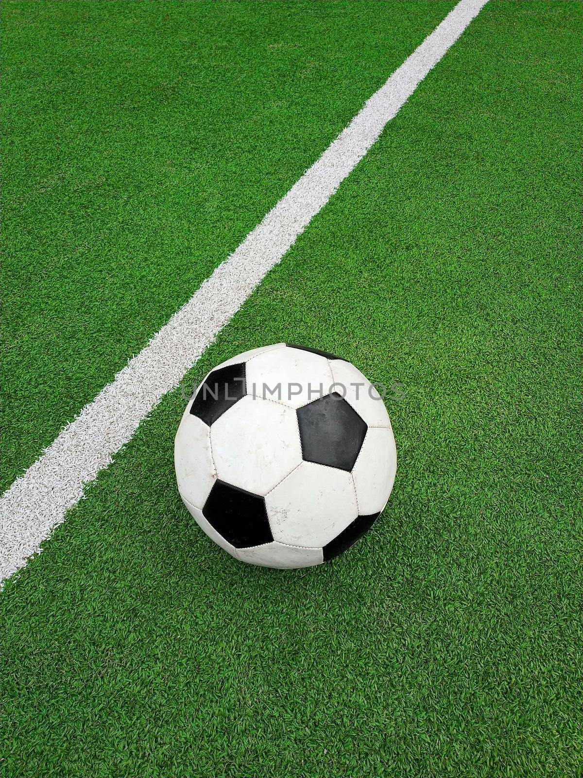 Close up one black and white football ball over green artificial turf of soccer field pitch with white marking lines, elevated, high angle view, personal perspective