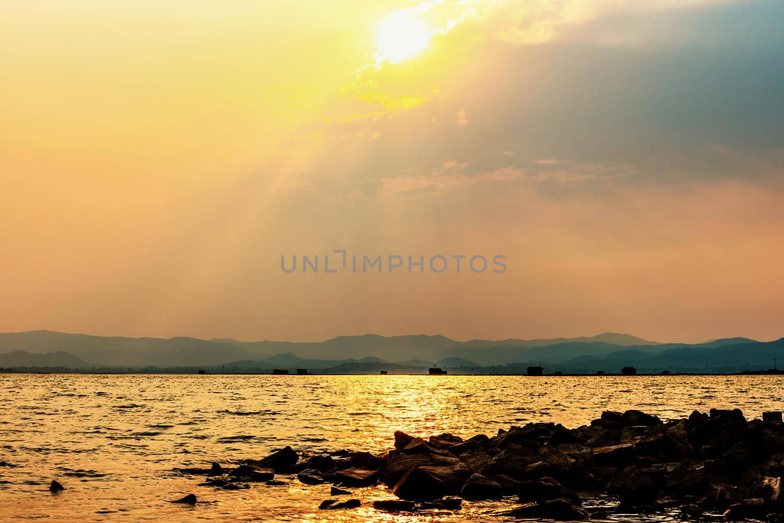 Beautiful nature landscape sun shines bright golden in the cloud reflect yellow light on the sky pile of rock in the water of a tropical lake mountain background at Krasiao Dam, Suphan Buri, Thailand