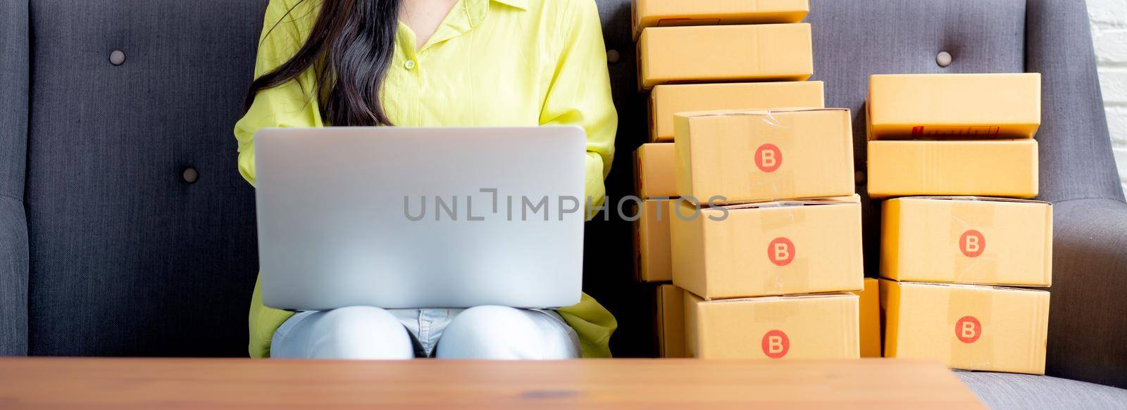 Young asian woman is merchant using laptop computer on sofa checking order of customer with purchase online shopping, business SME or startup, entrepreneur or seller and service delivery product. by nnudoo