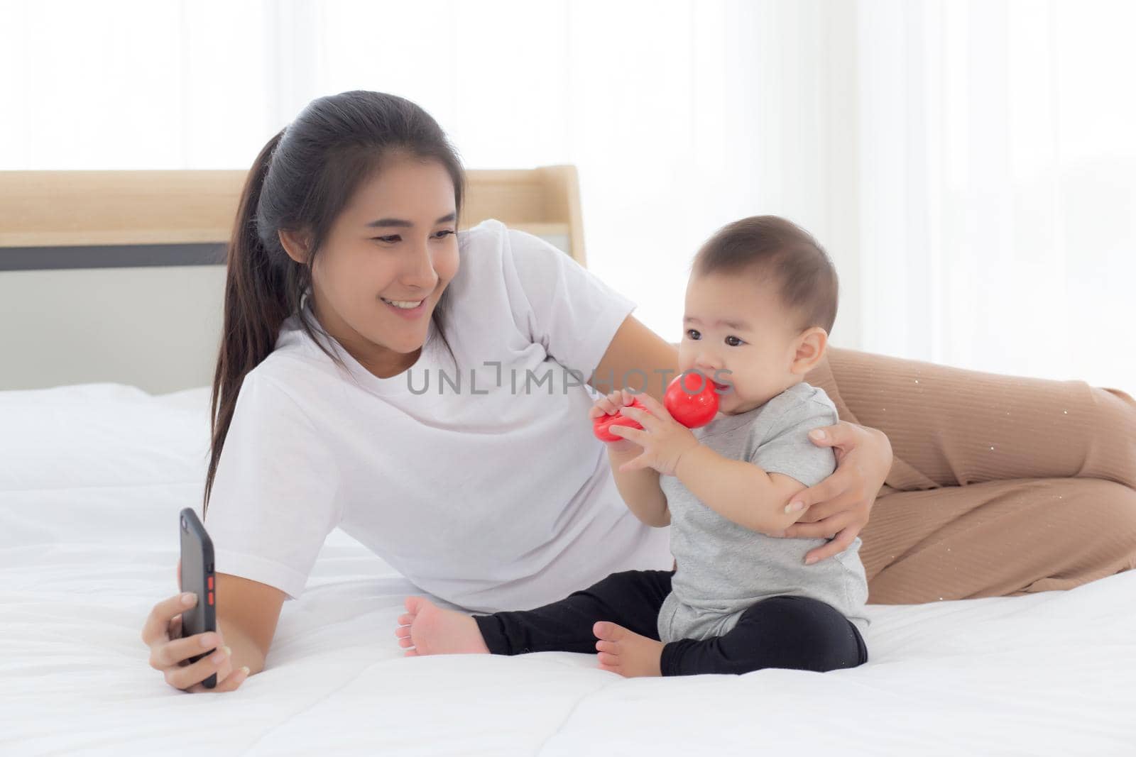 Young asian mother and little baby girl or newborn selfie with smart phone on bed in bedroom, happiness mom and daughter using phone video call at home, two people, family and communication concept.