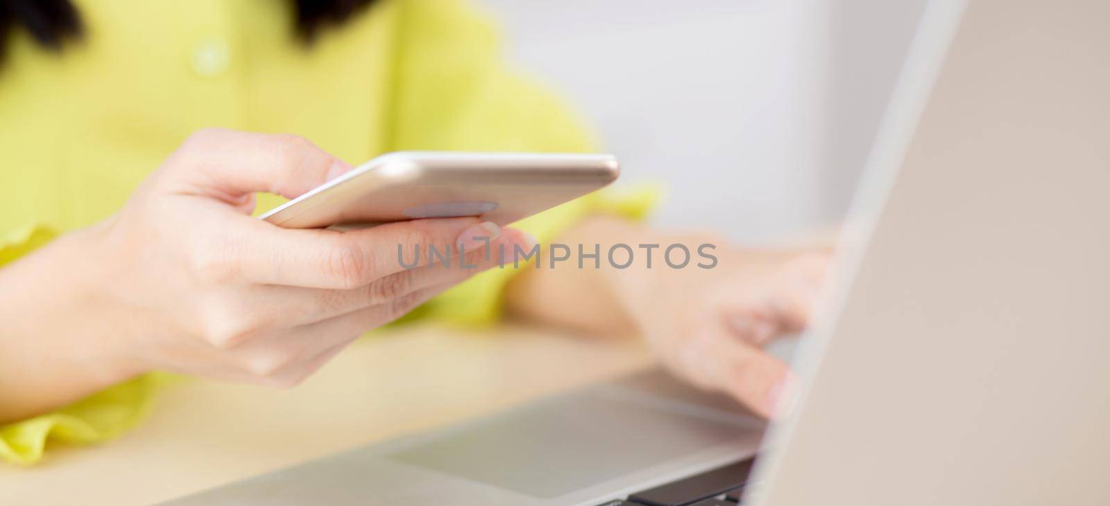 Closeup hand of young asian businesswoman working on laptop computer on desk at home office, freelance woman looking message on smart phone and typing on notebook, business and communication concept. by nnudoo