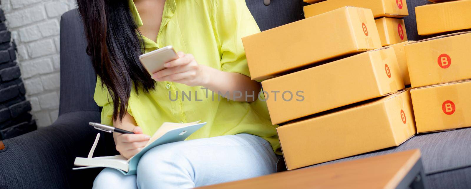 Young asian woman is entrepreneur looking phone and writing order of customer with online on notebook at home, online shopping, logistic and delivery, female is seller working on sofa, business SME. by nnudoo