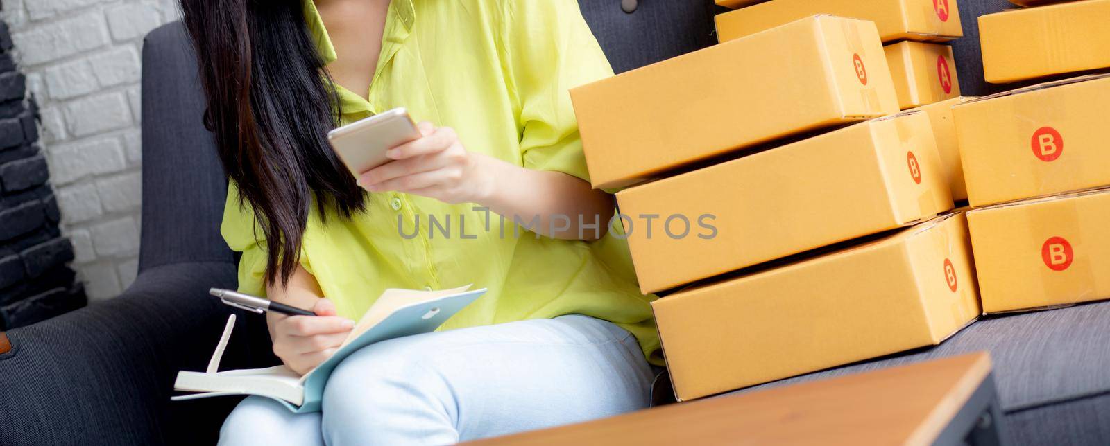 Young asian woman is entrepreneur looking phone and writing order of customer with online on notebook at home, online shopping, logistic and delivery, female is seller working on sofa, business SME.