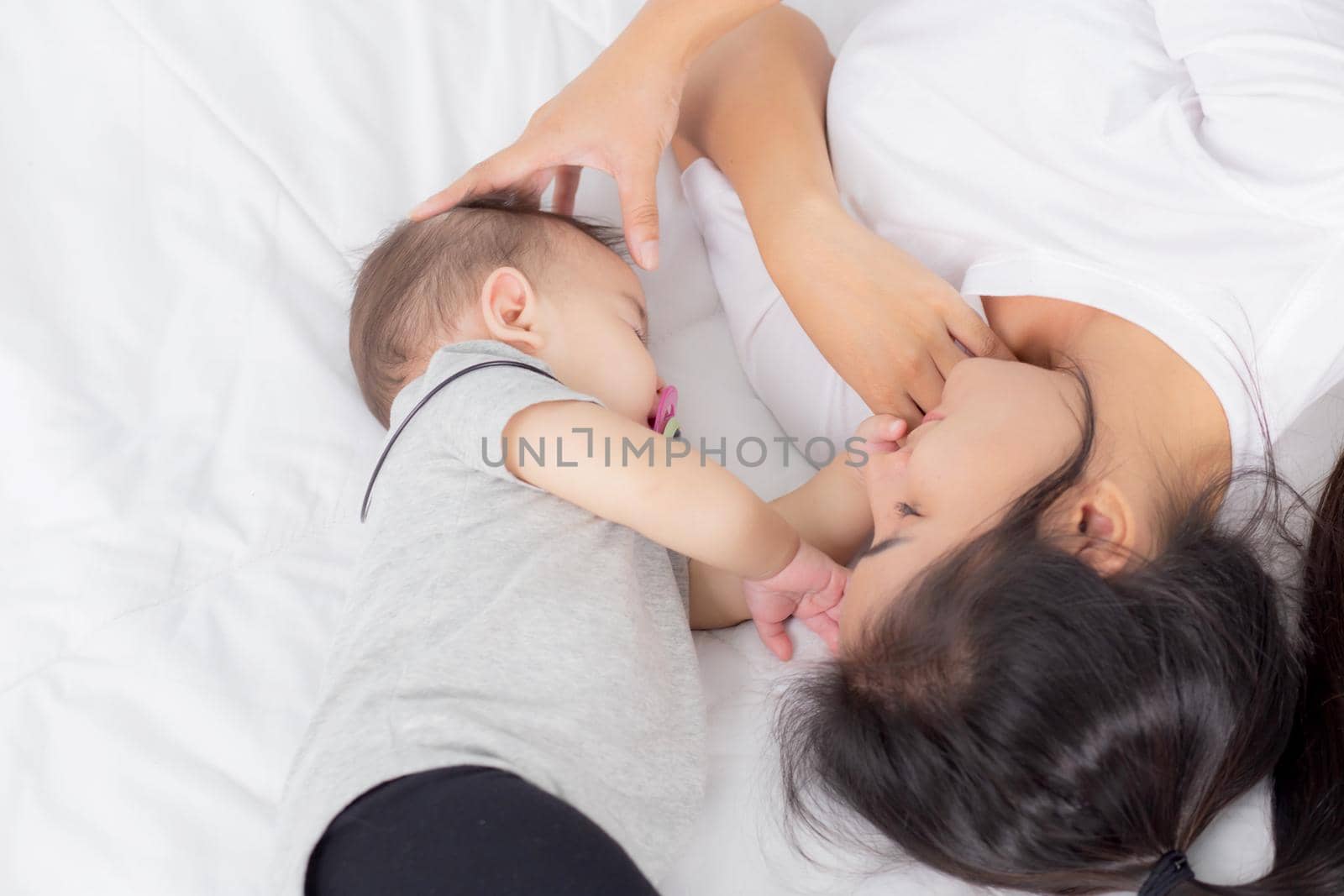 Young asian mother touch head of little baby girl with tender on bed in the bedroom, mom love newborn and care, woman with expression with child together, parent and daughter, family concept.