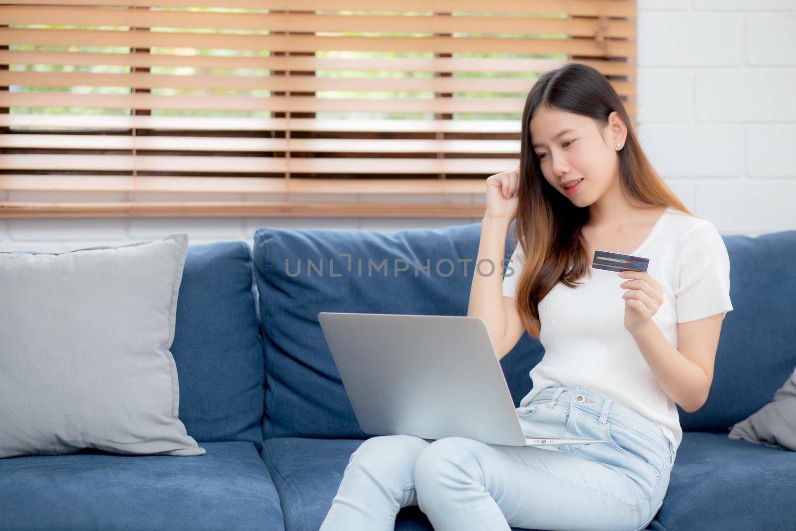 Young asian woman sit on sofa using laptop computer shopping online with credit card buying to internet, happy girl payment with e-business on couch, purchase and payment, business concept.