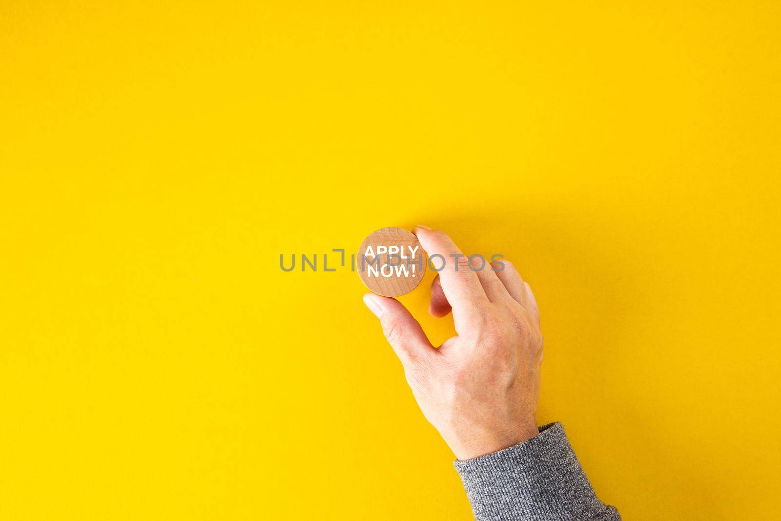 Male hand holds a wooden block with the word apply now on yellow background. Job vacancy, education or membership application concept.
