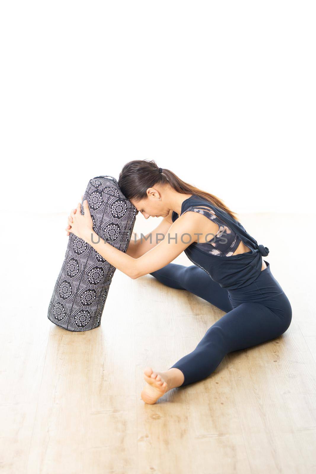 Restorative yoga with a bolster. Young sporty attractive woman in bright white yoga studio, lying on bolster cushion, stretching and relaxing during restorative yoga. Healthy active lifestyle.