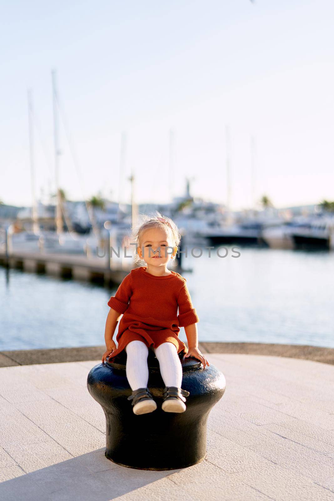 A portrait of a pretty 2-year old girl sitting on a boat pier by the sea. High quality photo