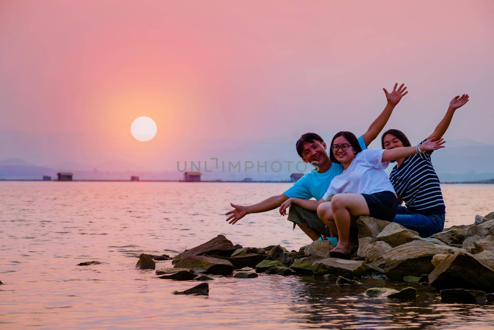 Asian family father mother and teenage daughter happy sitting on the waterfront at lake water sun in the orange sky beautiful nature landscape at sunset background, Krasiao Dam, Suphan Buri, Thailand