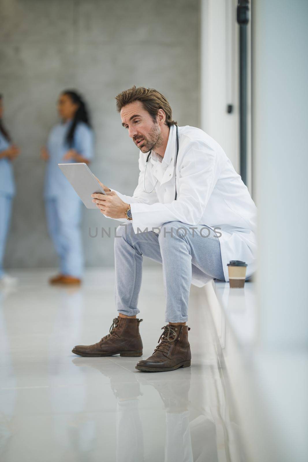 Shot of a mature doctor using digital tablet while sitting near a window in a hospital hallway during the Covid-19 pandemic.