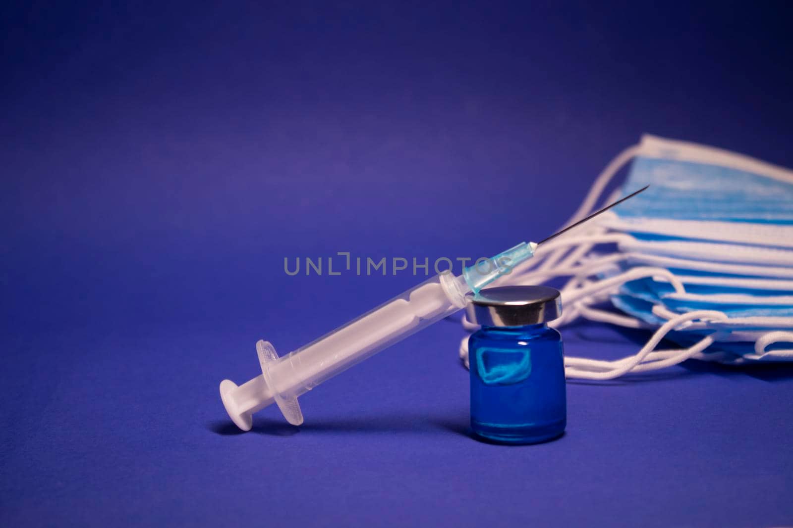 Vaccine bottle phial with no label and medical syringe with injection needle near pile of blue medical masks . isolated on blue background. cure. Development of coronavirus vaccine COVID19. copy space