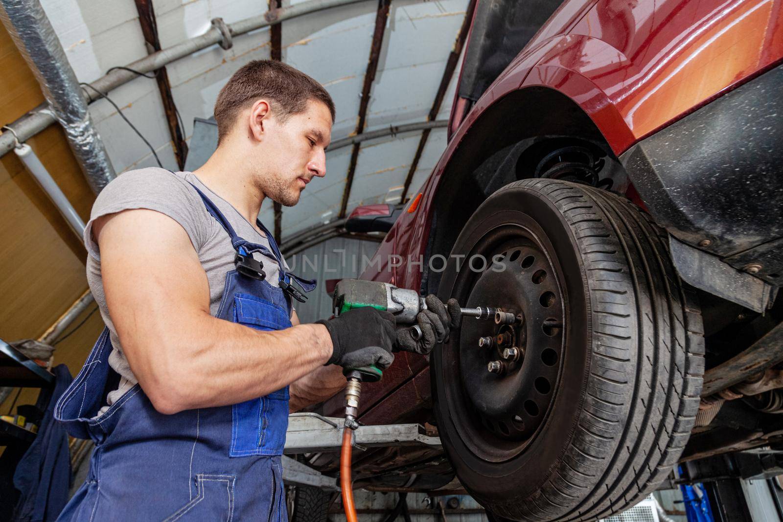 A muscular mechanic is maintaining wheels on a vehicle in auto garage with pneumatic tool.