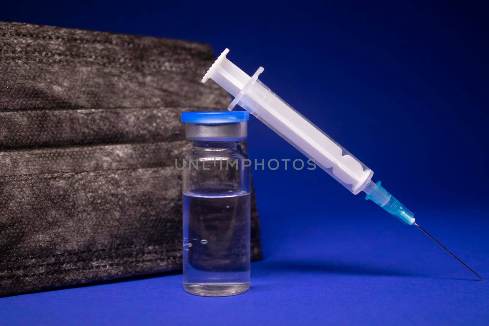 Vaccine bottle phial with no label near black medical mask and medical syringe with injection needle. isolated on blue background. cure. Development of coronavirus vaccine COVID-19. copy space