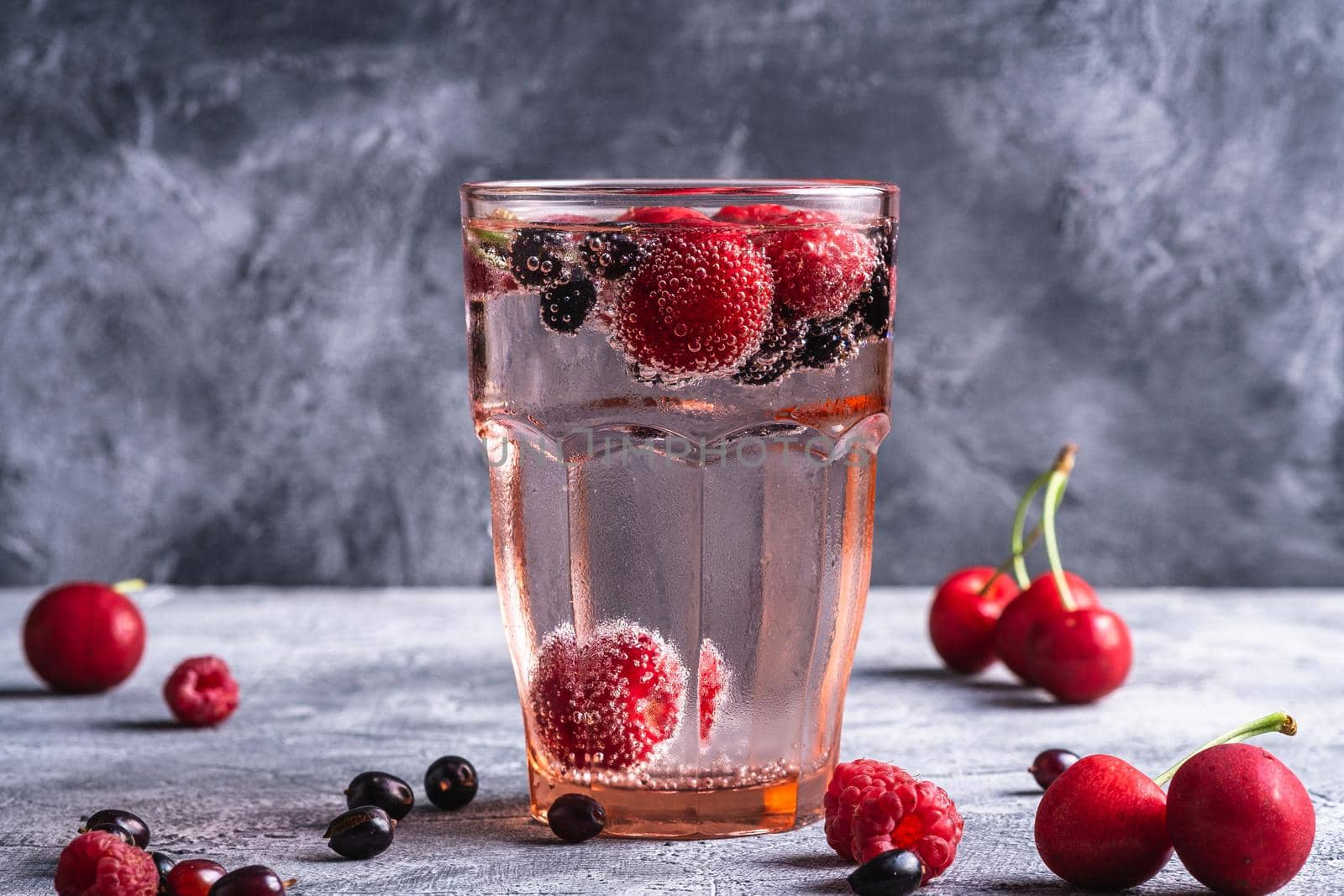 Fresh cold sparkling water drink with cherry, raspberry and currant berries in red faceted glass on stone concrete background, summer diet beverage, angle view