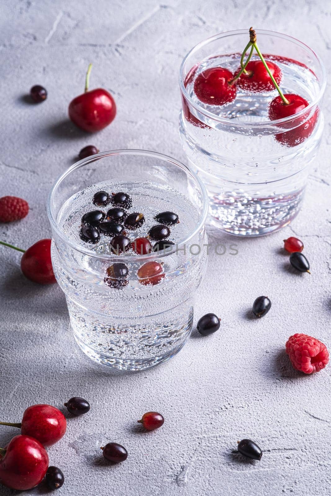 Fresh cold sparkling water drink with cherry, raspberry and currant berries in two transparent glass on stone concrete background, summer diet beverage, angle view macro by Frostroomhead