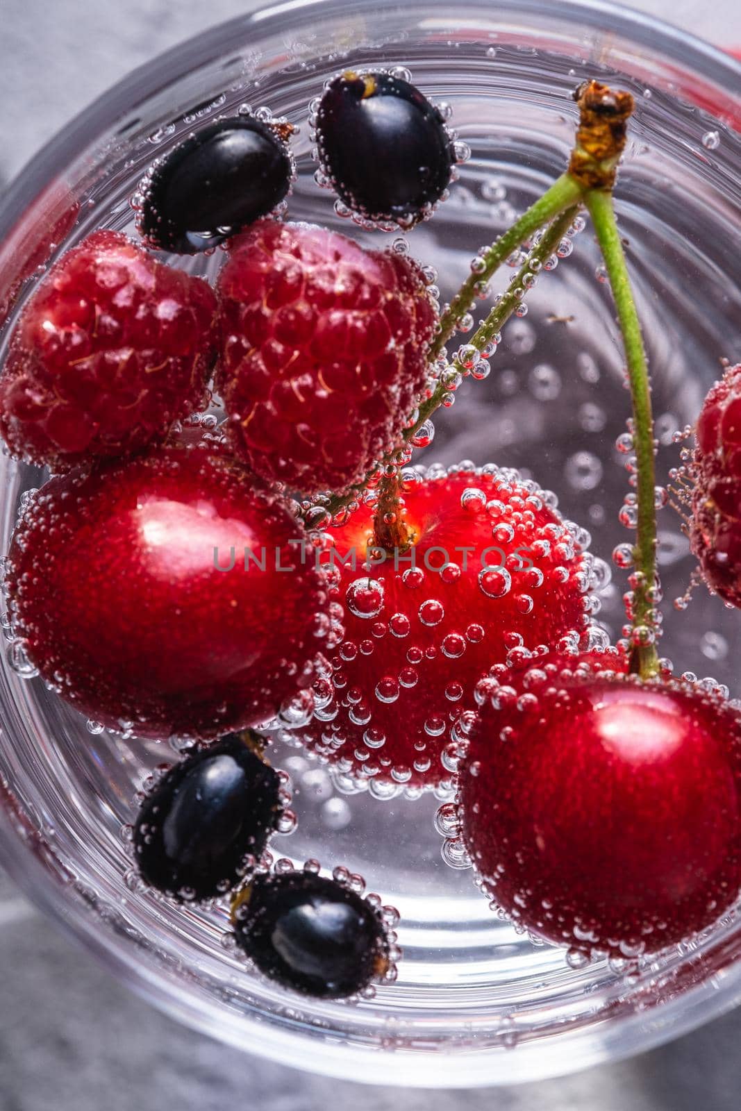 Fresh cold sparkling bubble water drink with cherry, raspberry and currant berries in transparent glass on stone concrete background, summer diet beverage, top view macro