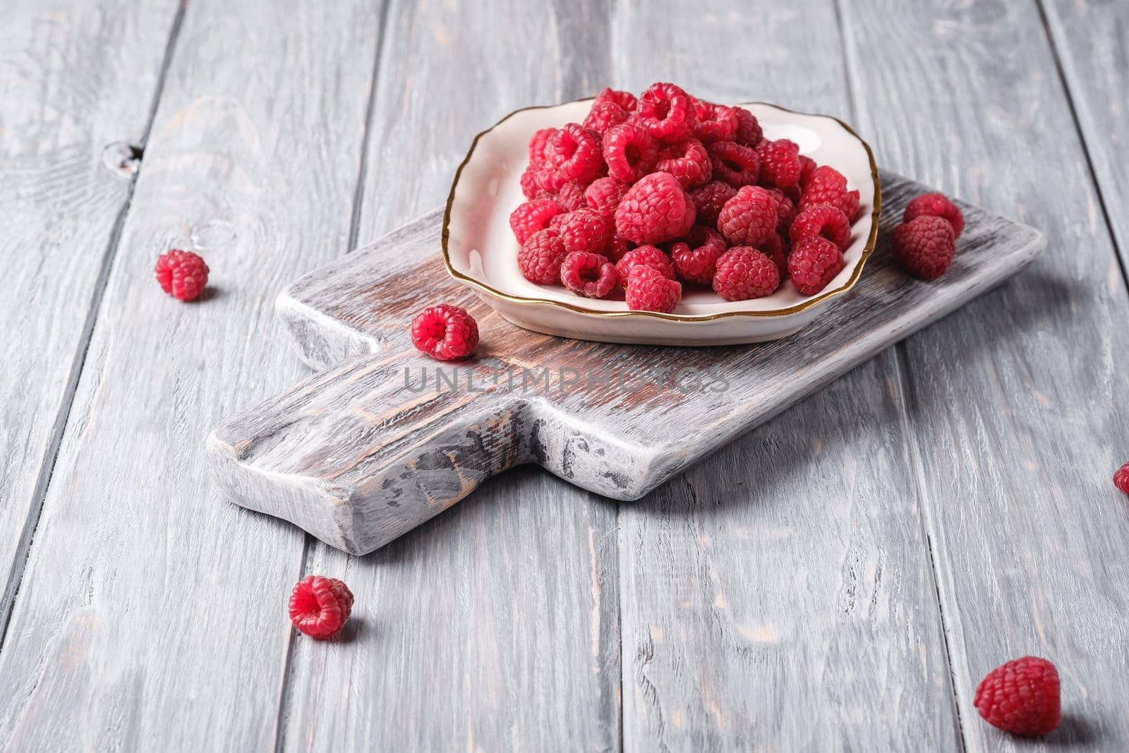 Raspberry fruits in plate on old cutting board, healthy pile of summer berries on grey wooden background, angle view