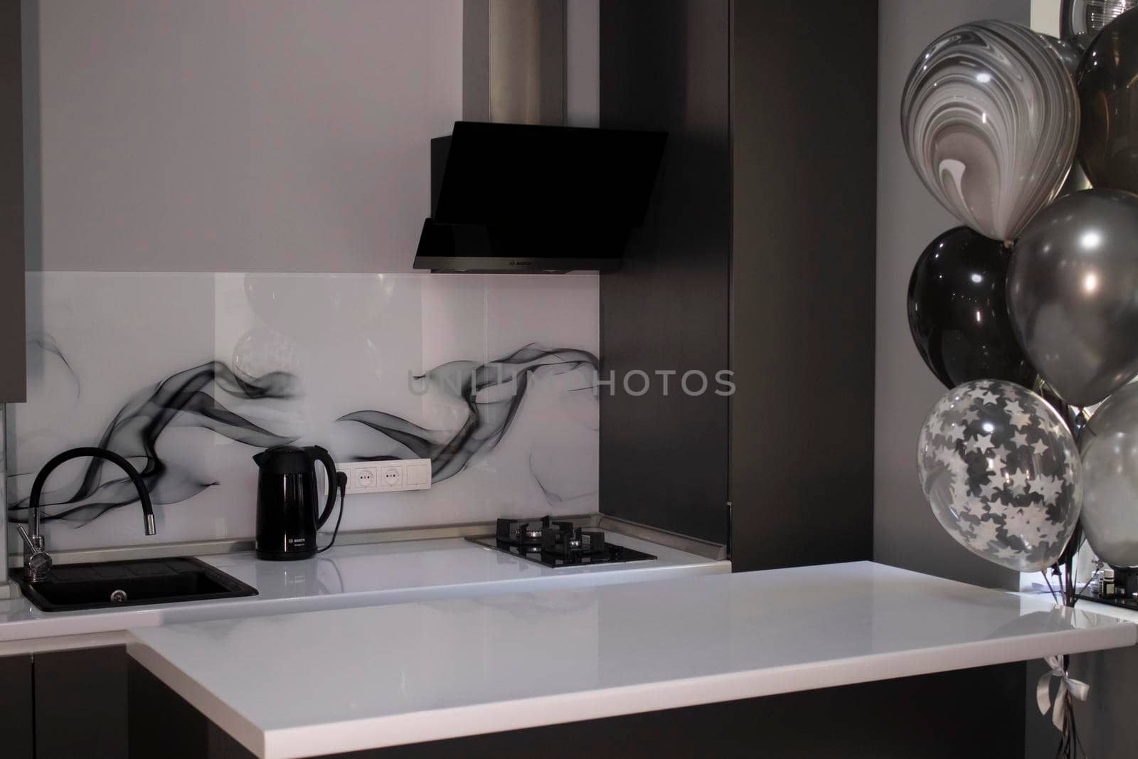 Corner of stylish Minimalistic grey kitchen, dark gray cupboards, white countertops and bar with stools. black sink, kettle and stove. and black and white balloons. High quality photo