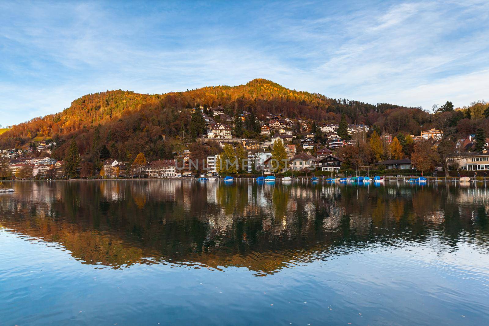 Idyllic panorama view of Thun town from Aare river side on sunny autumn day with colourful forest, Alps and blue sky cloud in background and beautiful reflection in water, Canton of Bern, Switzerland. by VogelSP